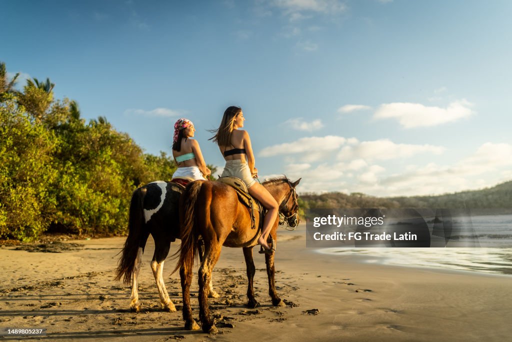 Amigas mirando hacia otro lado contemplando durante un paseo a caballo por la playa