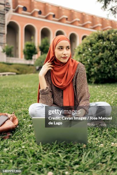 muslim woman siting on the grass using a computer - hiyab photos et images de collection