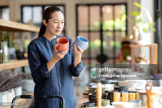 consumer decision-making process can help marketers understand the consumer's need. a japanese female shopping for home decor and necessities in a home decor store chooses a handmade ceramic mug between orange and blue colors from a shelf. - data retention stock pictures, royalty-free photos & images