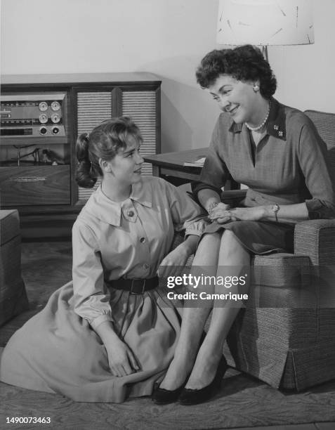 Well-dressed woman sitting in a chair in the living room. Her teenage daughter in a poodle skirt sits on the floor in front of her as they look at...