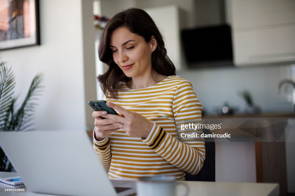 Young smiling businesswoman using a cellphone while working on a laptop in home office