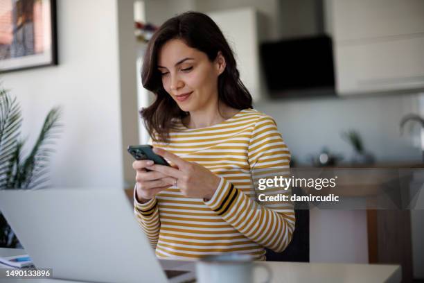 young smiling businesswoman using a cellphone while working on a laptop in home office - dispositivo de informação portátil imagens e fotografias de stock