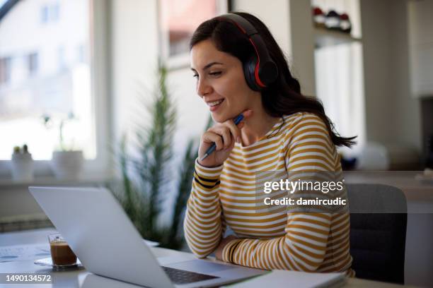 young woman with bluetooth headphones working on her laptop at home - zoom classroom stock pictures, royalty-free photos & images