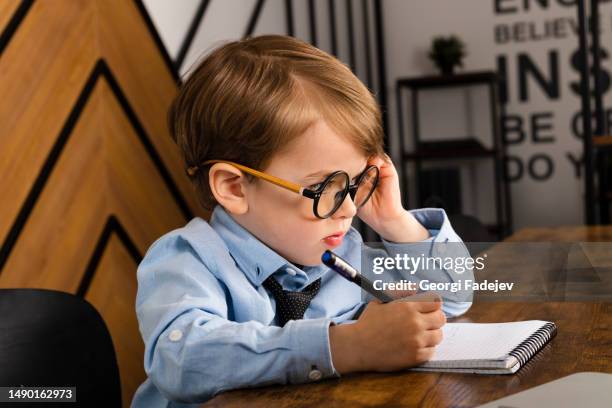 little boy in round eyeglasses, blue shirt and tie sitting at the desk in the office with a notepad. smart little kid, child prodigy. - child prodigy stock-fotos und bilder