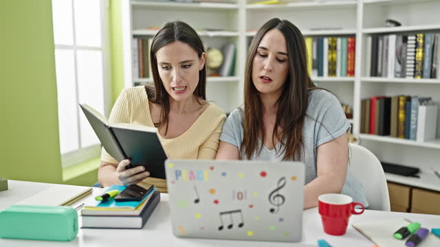 Two women sitting on table studying using laptop reading book at library university