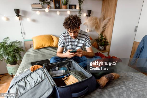 young man sitting on his bed, using smart phone and packing suitcase - arranjo imagens e fotografias de stock