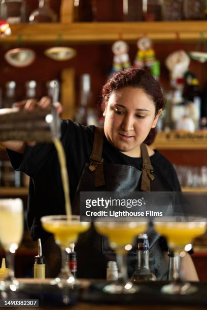 young adult bartender pours liquid from cocktail mixer into glasses for mango margaritas - barman tequila stockfoto's en -beelden