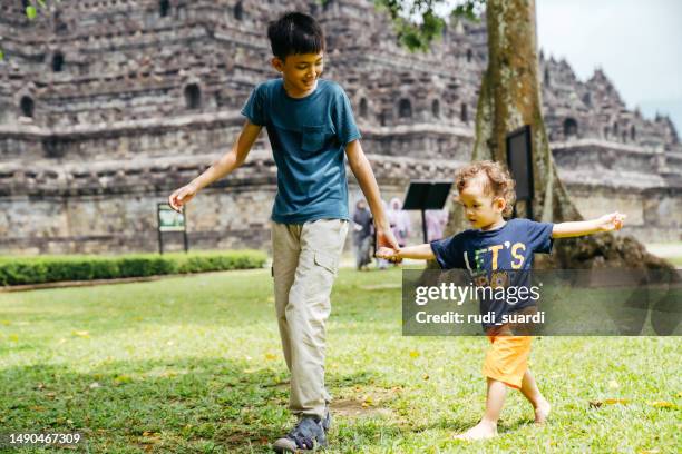 young kid with his little brother walking on the grass with borobudur temple in the background - old brother stock pictures, royalty-free photos & images