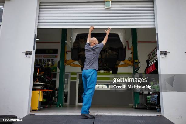 asian male mechanic opening shutter door to car workshop - autobergplaats stockfoto's en -beelden