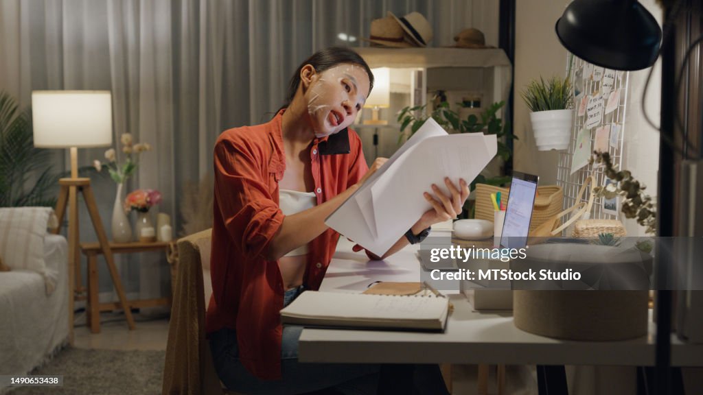 Une jeune femme asiatique utilise un masque assis devant le bureau avec un ordinateur portable et utilise un téléphone portable pour parler avec un collègue travaillant dans le salon de la maison la nuit. Travail à domicile.