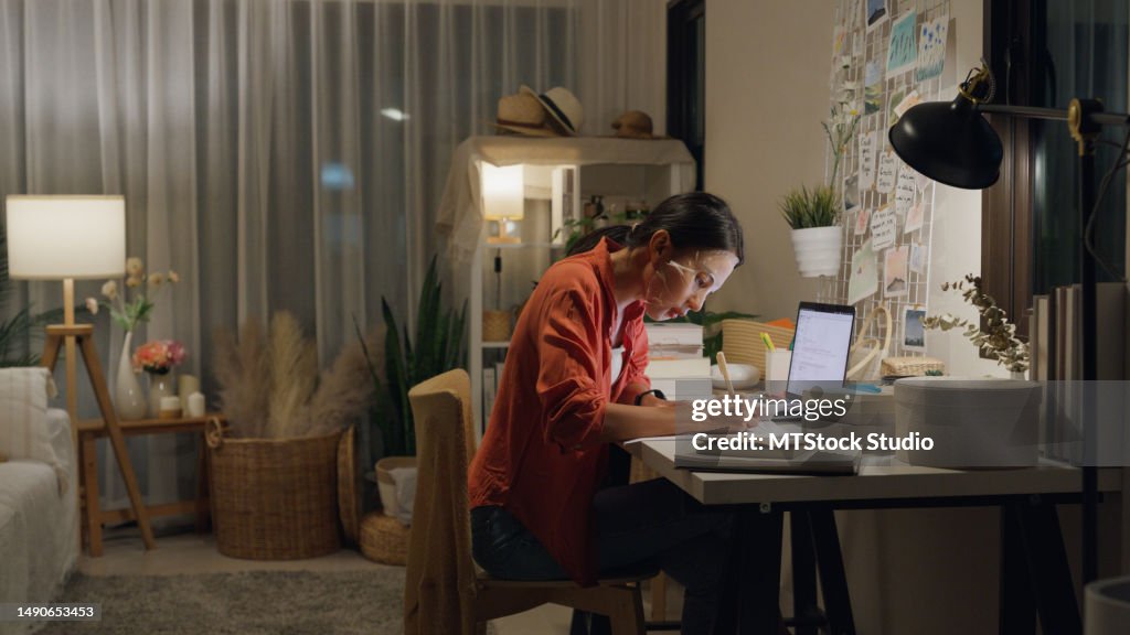 Young Asian woman use mask sheet on face sit front of desk with computer laptop and think create on work idea in living room at house at night. Work from home.