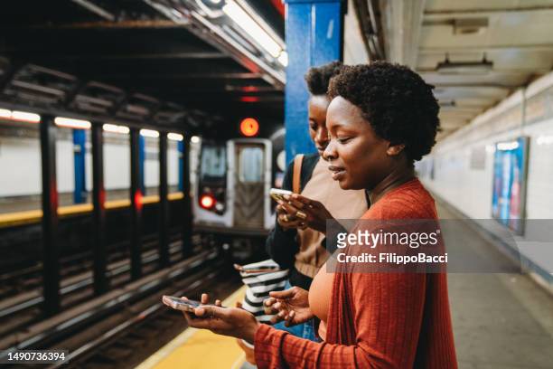 two friends are using smart phones in a subway station, waiting for the train - underground walkway stock pictures, royalty-free photos & images