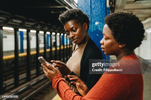 two friends are using smart phones in a subway station, waiting for the train - underground walkway stock pictures, royalty-free photos & images