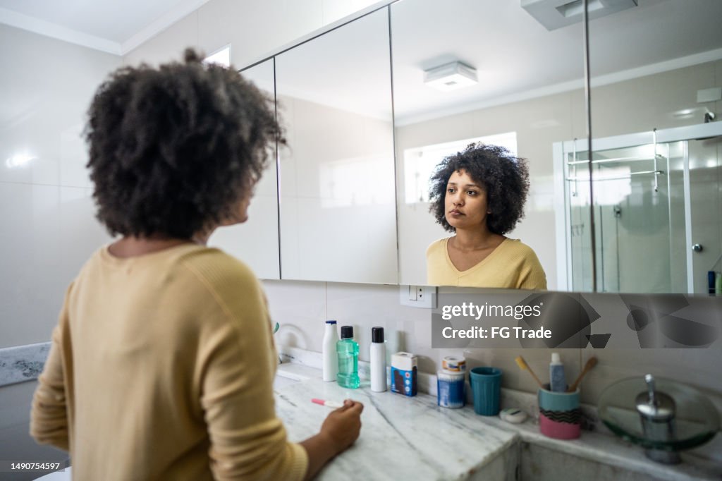 Sad young woman holding a pregnancy test looking at herself in mirror on bathroom at home