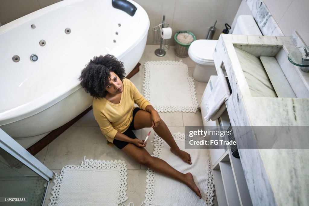 Sad young woman holding a pregnancy test sitting on ground in the bathroom at home