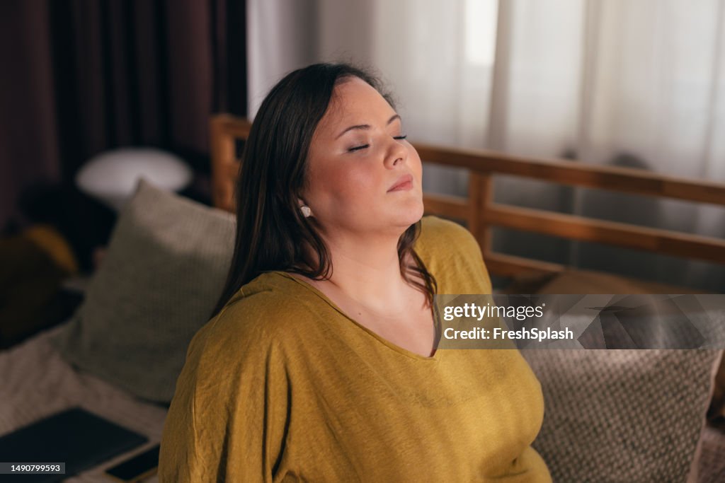 Relieving Stress and Anxiety: Beautiful Plus Size Woman Sitting on her Bed and Meditating with Eyes Closed