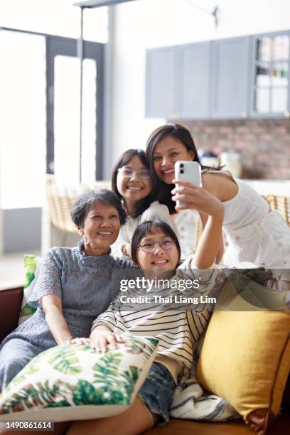 die enkelin benutzt ein smartphone, um ein selfie mit ihrer großmutter, ihrer mutter und ihrer jüngeren schwester zu machen. sie alle lächeln glücklich und veranschaulichen die schöne bindung innerhalb einer drei-generationen-familie. - chinese family taking photo at home stock-fotos und bilder