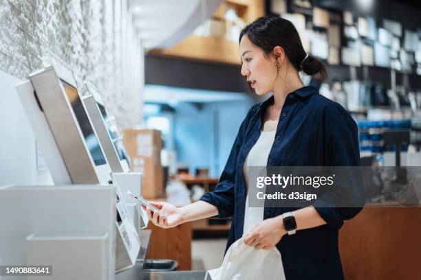 young asian woman with a reusable shopping bag, using contactless payment via smartphone to pay for her shopping at self-checkout kiosk in a store - authentication stock-fotos und bilder