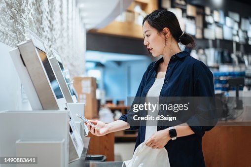 Young Asian woman with a reusable shopping bag, using contactless payment via smartphone to pay for her shopping at self-checkout kiosk in a store Young Asian woman with a reusable shopping bag, using contactless payment via smartphone to pay for her shopping at self-checkout kiosk in a store
