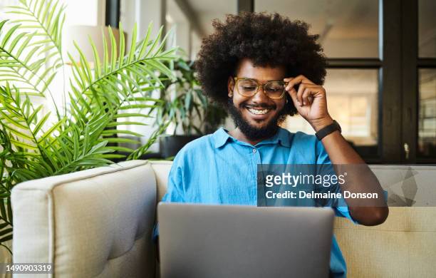 happy, young black man with an afro, sitting on the couch whilst looking at his laptop computer screen. - toothy smile stock pictures, royalty-free photos & images