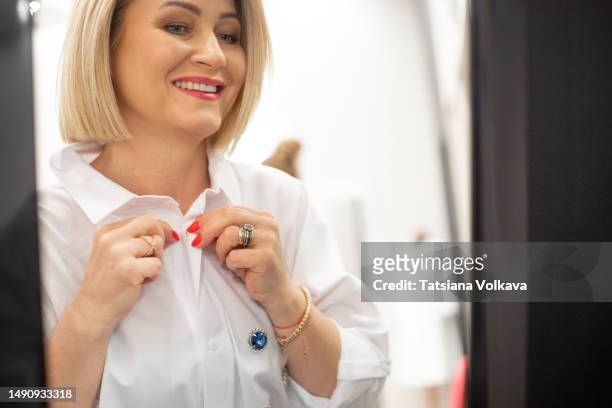 smiling mature business woman buttoning white shirt with blue brooch looking in mirror - abrochar fotografías e imágenes de stock