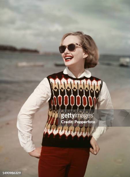 Vacation scene of a female fashion model posed wearing a sleeveless Fair Isle tank top sweater over a white blouse and burgundy coloured slacks, she...