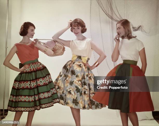 Posed studio portrait of three female fashion models wearing sleeveless and short sleeved tops and knee length full gathered skirts in printed...