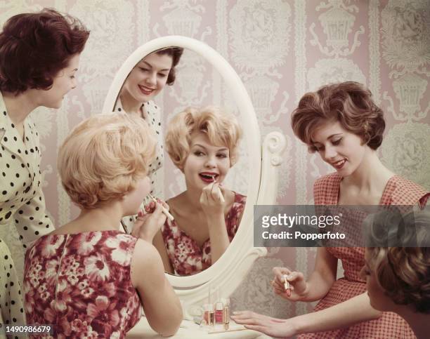 Posed studio portrait of three female fashion models gathered around a mirror and dressing table to watch fellow model Liz Duke demonstrating how she...