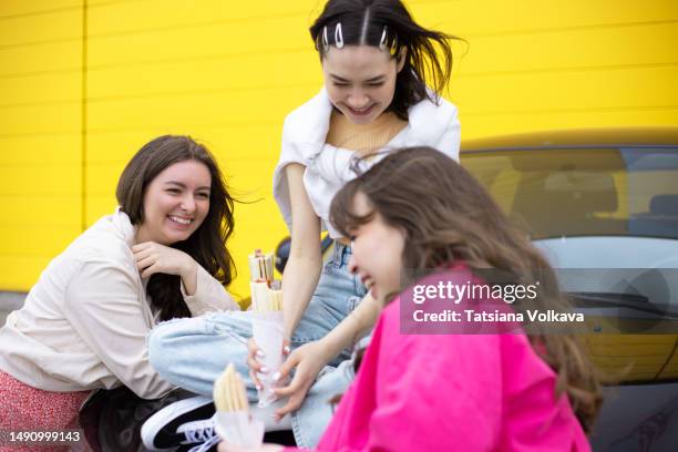 three female university students sitting on hood of black car by yellow wall eating hot dogs and laughing - mittagspause stock-fotos und bilder