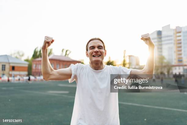 playful young man flexing his arms muscles with soccer field and bright sky in background - portrait playful caucasian man foto e immagini stock