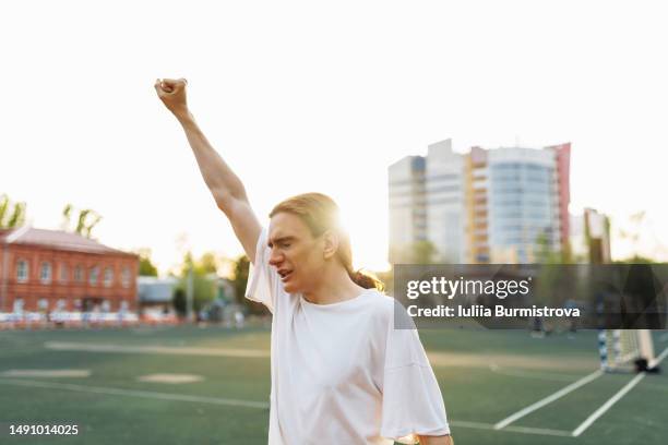 jubilant soccer fan standing with hand raised in triumph with bright sky and soccer field in background - siegerpose stock-fotos und bilder