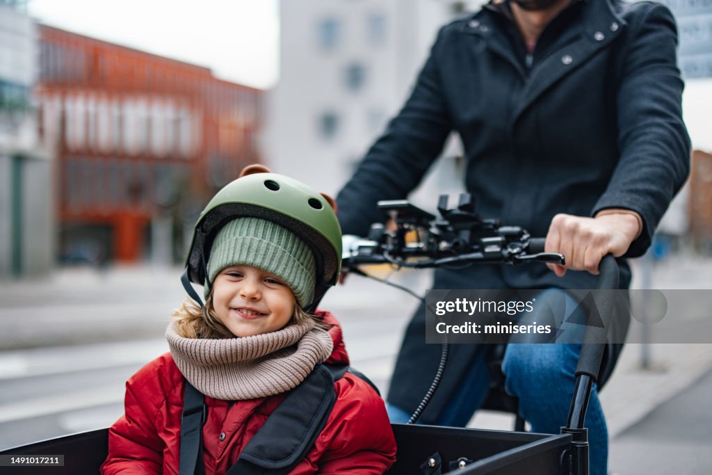 Unkenntlicher Vater, der mit seinem lächelnden kleinen Mädchen in der Stadt auf einem Lastenfahrrad fährt
