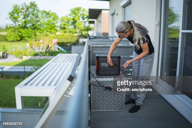 frau steckt kabel vom solarpanel in wiederaufladbares tragbares kraftwerk auf balkon ein - batteriespeicher stock-fotos und bilder