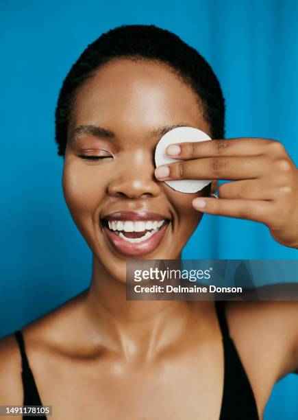 happy young black woman smiling whilst closed eyes holding one eye close with a cotton swap as she cleans her face and starts her facial routine, stock photo - cotton pad stock pictures, royalty-free photos & images