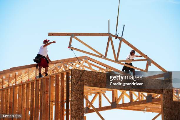 framing carpenters reaching to set a roof truss being lowered on to the top of a wall of a home being built on a sunny day - roof truss stock pictures, royalty-free photos & images