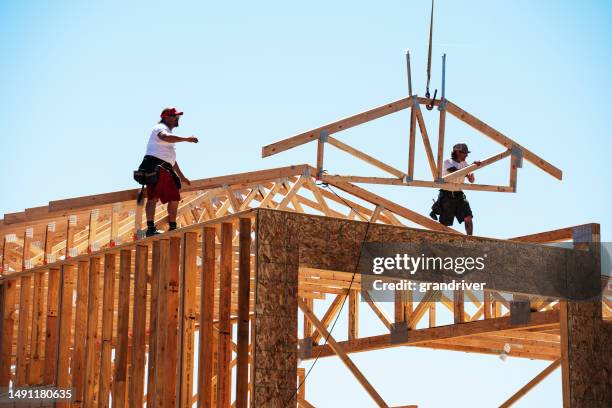 framing carpenters reaching to set a roof truss being lowered on to the top of a wall of a home being built on a sunny day - roof truss stock pictures, royalty-free photos & images