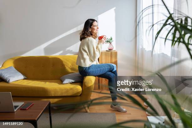 smiling young woman sitting on sofa while enjoying cup of coffee in the morning at home. domestic lifestyle concept - domestic room imagens e fotografias de stock