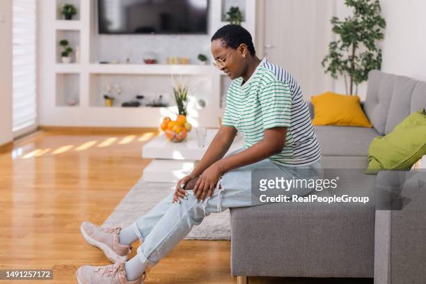 a young woman in pain holding her injured knee at home - injured knee stock pictures, royalty-free photos & images