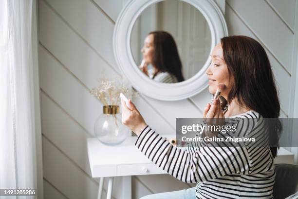 smiling brunette woman doing makeup with mobile phone online in bedroom at home - make up brush stock-fotos und bilder