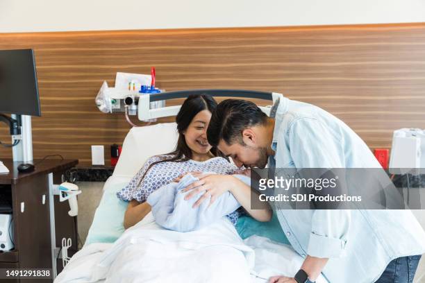 mid adult husband kisses newborn in wife's arms - family photo in the delivery room stock pictures, royalty-free photos & images