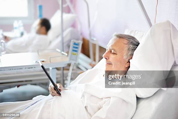 middle aged man reading a tablet in a hospital bed - postoperatief stockfoto's en -beelden