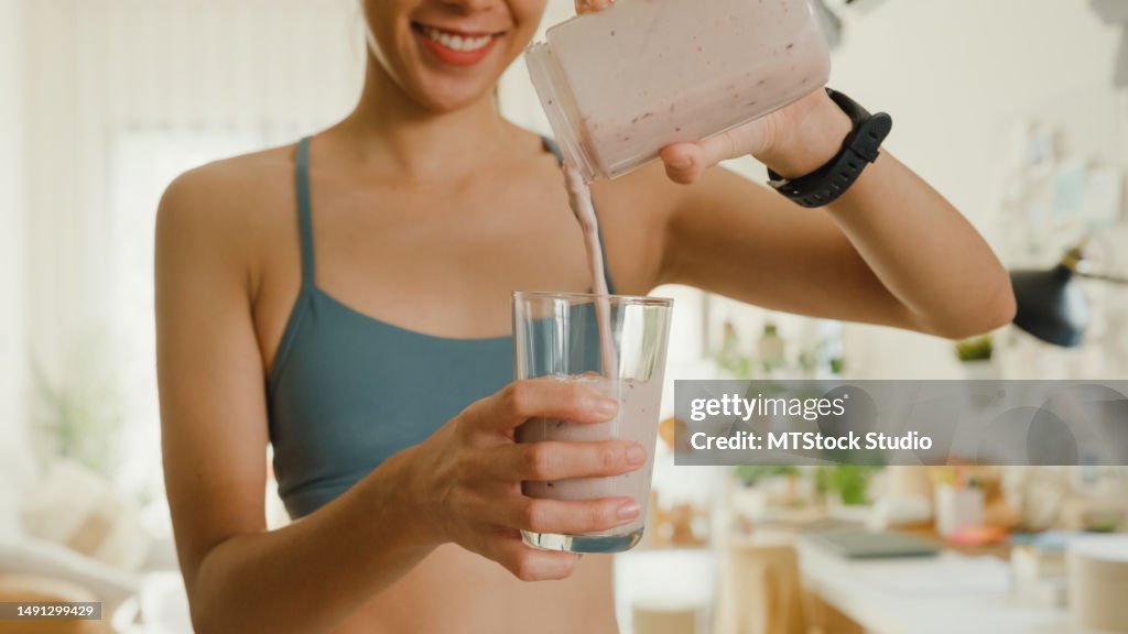 Closeup of young Asian athletic woman using blender and pouring smoothie to glass at home. Healthy food.