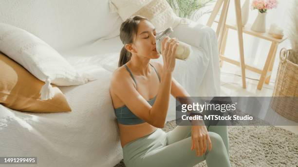 young asian athletic woman drinking protein shake in living room at home. diet and healthy food. - protein stockfoto's en -beelden