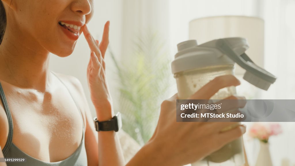 Closeup of young Asian athletic woman drinking protein shake on sofa in living room at home. Diet and healthy food.