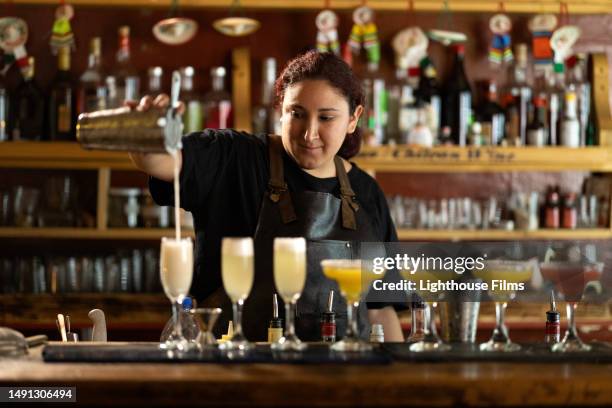 female bartender pours another mixed drink into glass next in line to colorful cocktails at bar - barman tequila stock-fotos und bilder