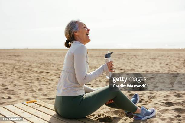 woman with water bottle sitting and laughing at beach - middle age active lifestyle stock pictures, royalty-free photos & images