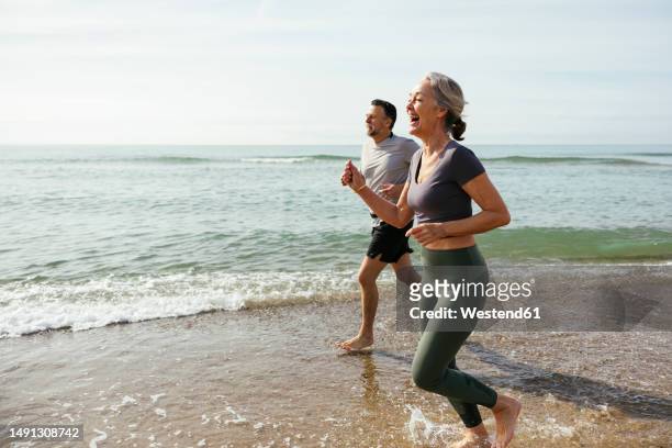 happy mature couple laughing and running in water at beach - mature woman in water stock pictures, royalty-free photos & images