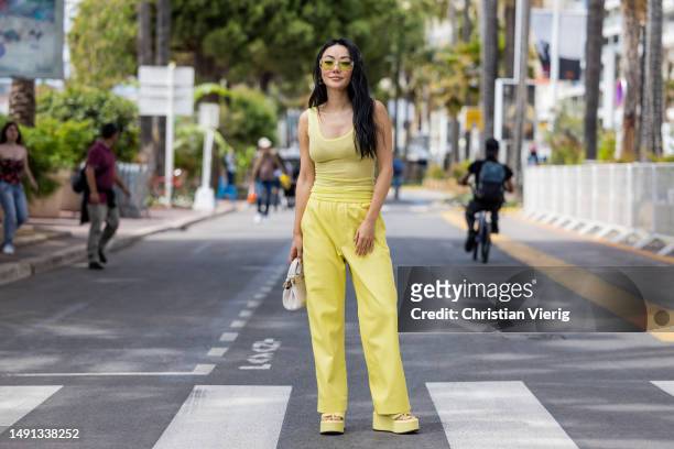 Jessica Wang wears yellow pants, shirt, sunglasses, white bag, platform sandals during Cannes Film Festival on May 18, 2023 in Cannes, France.