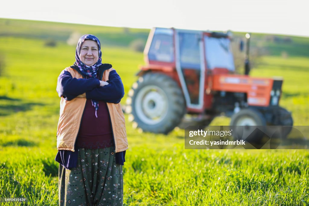 Senior farmer woman with his arms tied standing in front of his orange tractor