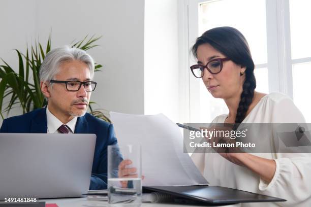 middle shot and frontal view of a japanese senior businessman and beautiful ceo businesswoman sitting on a meeting table near a window using a laptop and looking at documents and talking about new agreements at the workplace. - laptop frontal stock pictures, royalty-free photos & images